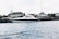 A sleek, modern yacht is moored along a waterfront with a historic building featuring a green copper dome in the background. The scene is set against a lightly clouded sky and the water appears calm with small ripples.