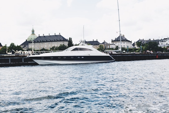 A sleek, modern yacht is moored along a waterfront with a historic building featuring a green copper dome in the background. The scene is set against a lightly clouded sky and the water appears calm with small ripples.