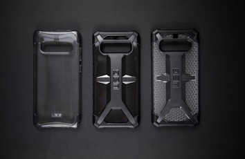 Three protective phone cases are lined up against a dark background. The cases feature rugged designs with one being clear and the others displaying a distinct cross pattern with textured surfaces.