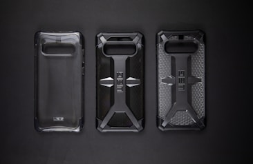 Three protective phone cases are lined up against a dark background. The cases feature rugged designs with one being clear and the others displaying a distinct cross pattern with textured surfaces.