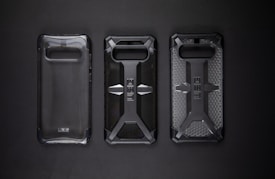 Three protective phone cases are lined up against a dark background. The cases feature rugged designs with one being clear and the others displaying a distinct cross pattern with textured surfaces.