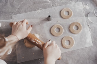Hands using a rolling pin to flatten dough on a surface, surrounded by circular dough cutouts and a pastry tip. The scene captures a baking process with ingredients and tools visible.
