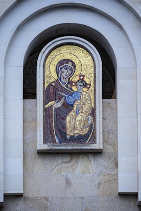 A mosaic artwork featuring a religious icon embedded in a stone wall. The figures depicted are dressed in traditional garments, with intricate designs and a backdrop of shimmering gold tiles.