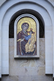 A mosaic artwork featuring a religious icon embedded in a stone wall. The figures depicted are dressed in traditional garments, with intricate designs and a backdrop of shimmering gold tiles.