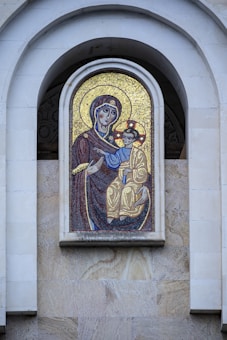 A mosaic artwork featuring a religious icon embedded in a stone wall. The figures depicted are dressed in traditional garments, with intricate designs and a backdrop of shimmering gold tiles.