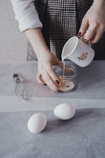 A person prepares ingredients on a kitchen counter, pouring a liquid from a decorative mug into a clear glass bowl. Nearby, two white eggs rest on the parchment paper next to a metal whisk.