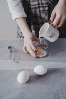 A person prepares ingredients on a kitchen counter, pouring a liquid from a decorative mug into a clear glass bowl. Nearby, two white eggs rest on the parchment paper next to a metal whisk.
