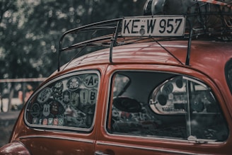 A vintage red car with a classic, rounded shape features a window covered in various stickers. The roof rack holds a spare tire and other items. The scene is set outdoors with a blurred background of trees.