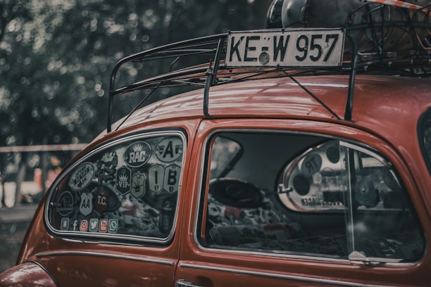 A vintage red car with a classic, rounded shape features a window covered in various stickers. The roof rack holds a spare tire and other items. The scene is set outdoors with a blurred background of trees.