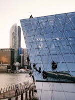 Several workers are suspended on ropes, performing maintenance or cleaning on the glass exterior of a modern, angular building by the waterfront. The building features a geometric, grid-like design and reflects the surrounding urban skyline, including high-rise buildings. A person is walking on a curving boardwalk below.