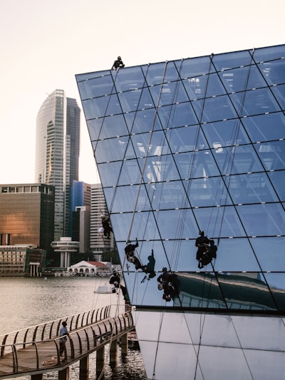Several workers are suspended on ropes, performing maintenance or cleaning on the glass exterior of a modern, angular building by the waterfront. The building features a geometric, grid-like design and reflects the surrounding urban skyline, including high-rise buildings. A person is walking on a curving boardwalk below.
