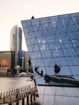 Several workers are suspended on ropes, performing maintenance or cleaning on the glass exterior of a modern, angular building by the waterfront. The building features a geometric, grid-like design and reflects the surrounding urban skyline, including high-rise buildings. A person is walking on a curving boardwalk below.