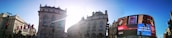 Urban scene featuring historical buildings with ornate architecture, a curved digital billboard displaying an advertisement, and a bright, sunny sky with the sun's rays between the buildings.