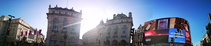 Urban scene featuring historical buildings with ornate architecture, a curved digital billboard displaying an advertisement, and a bright, sunny sky with the sun's rays between the buildings.