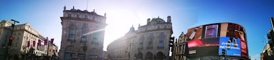 Urban scene featuring historical buildings with ornate architecture, a curved digital billboard displaying an advertisement, and a bright, sunny sky with the sun's rays between the buildings.