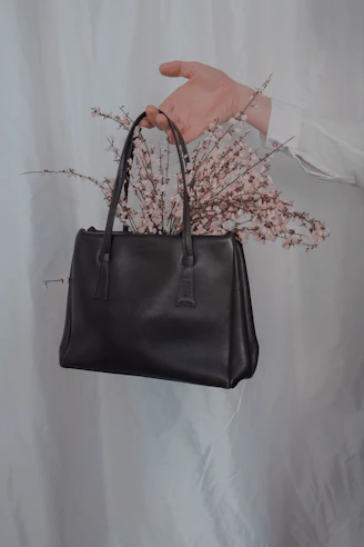 A sleek, minimalist handbag resting on a soft, neutral-toned fabric with delicate cherry blossom petals scattered around.