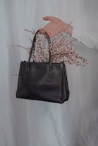 A hand emerges from a light-colored background, holding a black leather handbag. Delicate pink flowers appear to be arranged inside the handbag, creating an elegant and artistic composition.