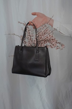 A hand emerges from a light-colored background, holding a black leather handbag. Delicate pink flowers appear to be arranged inside the handbag, creating an elegant and artistic composition.