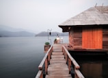 A wooden cabin with a slanted roof is situated by a tranquil lake. A wooden dock extends from the cabin towards the water, with a few potted plants and a single outdoor chair placed at the end of the dock. Misty hills and trees can be seen in the background, creating a serene and peaceful setting.