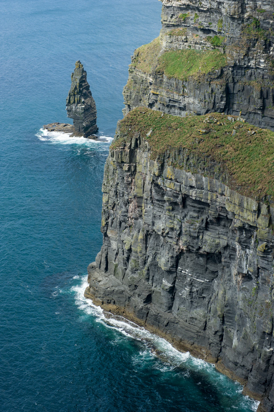 The image depicts a dramatic cliff face with two distinct features - a tall, pointed rock formation on the left side and a smaller, rounded rock formation on the right side. The cliff face is covered in green vegetation, adding a touch of color to the otherwise gray landscape. The deep blue ocean forms the backdrop for this rugged natural scene.