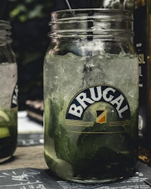A close-up view of a glass jar filled with a cold beverage, containing ice cubes and mint leaves. The jar is branded with a logo that reads 'Brugal.' Two black straws are placed inside. The background is blurred, featuring another similar jar and some green foliage.