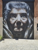 A large, detailed mural of an elderly person's face painted on a garage door. The artwork captures deep wrinkles and expressive eyes, highlighting the features with shades of black, white, and gray. The cobblestone ground leads up to the mural, which is set against a rustic wall.