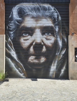 A large, detailed mural of an elderly person's face painted on a garage door. The artwork captures deep wrinkles and expressive eyes, highlighting the features with shades of black, white, and gray. The cobblestone ground leads up to the mural, which is set against a rustic wall.
