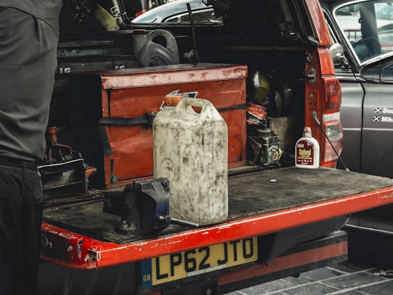 A red utility vehicle has its tailgate lowered to reveal the back section filled with various tools and containers. Prominent items include a large white plastic canister sitting on the surface, a red toolbox, a smaller metal can labeled 'Solvol,' and a blue metal vise attached to the tailgate. A portion of a person's body is visible on the left, suggesting a workshop or repair setting.