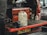A red utility vehicle has its tailgate lowered to reveal the back section filled with various tools and containers. Prominent items include a large white plastic canister sitting on the surface, a red toolbox, a smaller metal can labeled 'Solvol,' and a blue metal vise attached to the tailgate. A portion of a person's body is visible on the left, suggesting a workshop or repair setting.