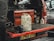 A red utility vehicle has its tailgate lowered to reveal the back section filled with various tools and containers. Prominent items include a large white plastic canister sitting on the surface, a red toolbox, a smaller metal can labeled 'Solvol,' and a blue metal vise attached to the tailgate. A portion of a person's body is visible on the left, suggesting a workshop or repair setting.