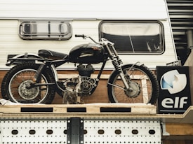 A vintage motorcycle is displayed in front of a white camper. The motorcycle has a classic design with a black frame, tank, and fenders. A sign with the logo and name 'elf' is situated below, partially visible. The setting appears industrial or workshop-like, with a metal and wood backdrop.