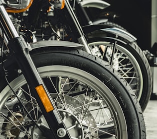 Close-up view of several motorcycle wheels lined up, featuring intricate spokes and prominent tire treads. The focus is on the details of the front tires and fenders, with a shiny metallic finish and visible disc brakes.