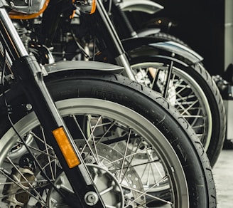 Close-up view of several motorcycle wheels lined up, featuring intricate spokes and prominent tire treads. The focus is on the details of the front tires and fenders, with a shiny metallic finish and visible disc brakes.
