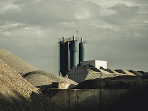 A construction or industrial site with piles of gravel or rocks surrounding silos and industrial buildings. The sky is overcast, contributing a muted gray tone to the setting.