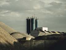 A construction or industrial site with piles of gravel or rocks surrounding silos and industrial buildings. The sky is overcast, contributing a muted gray tone to the setting.