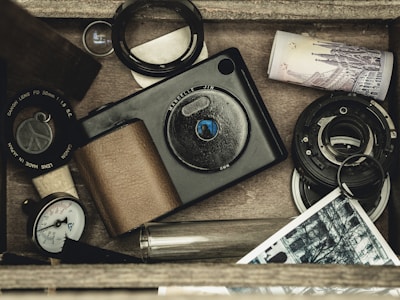 A collection of vintage photography equipment placed in a wooden box. There is a camera, a camera lens, photo slides, a roll of film, a currency note, a lens hood, and a small dial gauge. The items are arranged in an orderly, but casual manner giving a sense of nostalgia.