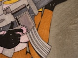 A stylized painting depicting a hand holding a rifle, with a focus on the magazine and trigger area. The artwork features bold outlines and a simple palette, creating a graphic novel or pop art appearance.