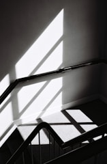 An abstract black and white photograph featuring a staircase with geometric shadows and highlights. The image has a minimalist aesthetic with strong diagonal lines and contrasts between light and shadow.