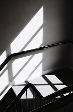 An abstract black and white photograph featuring a staircase with geometric shadows and highlights. The image has a minimalist aesthetic with strong diagonal lines and contrasts between light and shadow.