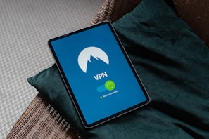 A tablet displaying a VPN app is placed on a green cushion on a wicker chair. The screen shows the VPN logo on a blue background with a green toggle indicating that the connection is active.