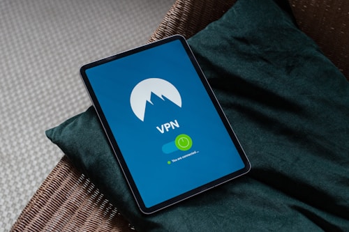 A tablet displaying a VPN app is placed on a green cushion on a wicker chair. The screen shows the VPN logo on a blue background with a green toggle indicating that the connection is active.