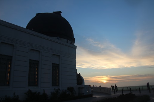 A large domed building, likely an observatory, is silhouetted against a sunset. The sky is filled with soft clouds and warm hues of orange and blue. Several people are visible on a walkway, gazing towards the setting sun, creating a peaceful ambiance.