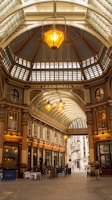 An ornate indoor marketplace with a high, arched glass roof and detailed architectural elements. Warm lighting from large hanging lamps creates a welcoming atmosphere. The building's name, 'Leadenhall', is prominently displayed. Shops with traditional signage line the sides, and a few people are visible walking below.