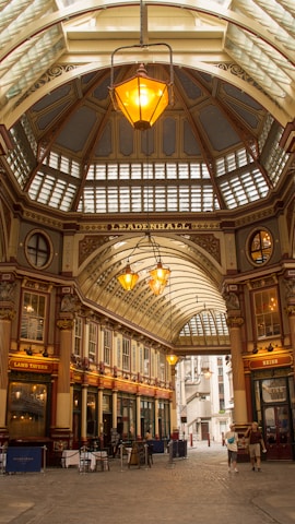 An ornate indoor marketplace with a high, arched glass roof and detailed architectural elements. Warm lighting from large hanging lamps creates a welcoming atmosphere. The building's name, 'Leadenhall', is prominently displayed. Shops with traditional signage line the sides, and a few people are visible walking below.