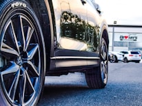 A close-up of a car's tire and wheel on a paved surface, reflecting sunlight. In the background, a car dealership with modern architecture and some parked vehicles are visible.