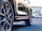 A close-up of a car's tire and wheel on a paved surface, reflecting sunlight. In the background, a car dealership with modern architecture and some parked vehicles are visible.