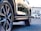 A close-up of a car's tire and wheel on a paved surface, reflecting sunlight. In the background, a car dealership with modern architecture and some parked vehicles are visible.