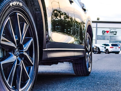 A close-up of a car's tire and wheel on a paved surface, reflecting sunlight. In the background, a car dealership with modern architecture and some parked vehicles are visible.