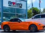 A bright orange convertible sports car is parked outside a car dealership. The building has a sign with 'Brown's FAIRFAX MAZDA' on it, and there are two flags flying nearby. A larger white SUV is also parked next to the sports car. Trees and a clear blue sky provide a backdrop.