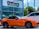 A bright orange convertible sports car is parked outside a car dealership. The building has a sign with 'Brown's FAIRFAX MAZDA' on it, and there are two flags flying nearby. A larger white SUV is also parked next to the sports car. Trees and a clear blue sky provide a backdrop.